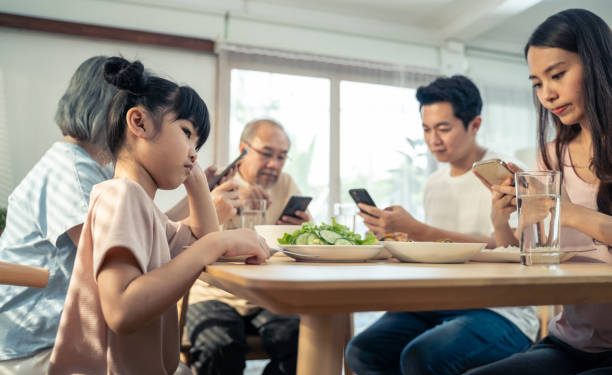Upset daughter sitting at lunch table looking to addicted parents and grandparents using smartphones while eating breakfast. Ignoring Family not paying attention on small child. bad parenting in house